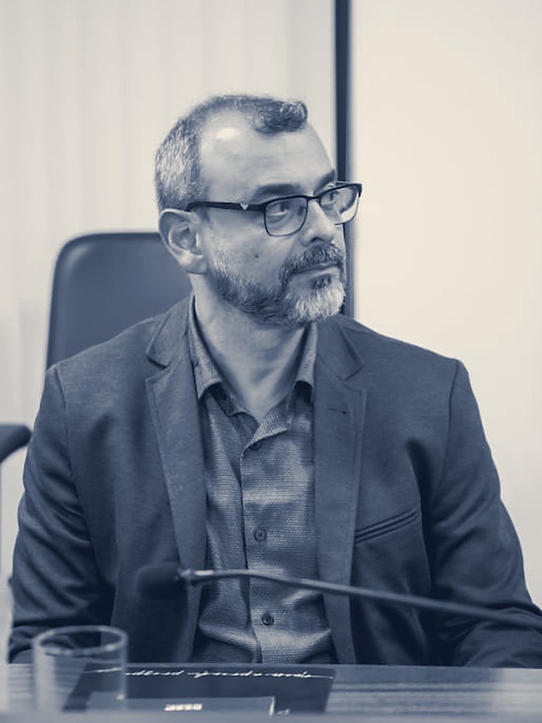 Guilherme Corrêa sentado à mesa do lançamento do livro, de paletó cinza, com um microfone à frente